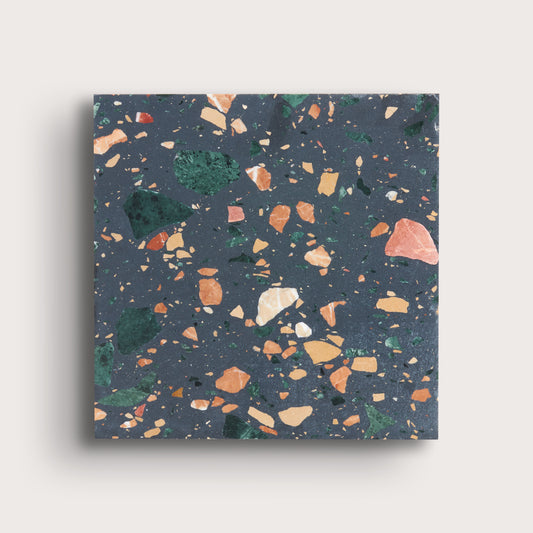 A square terrazzo-patterned tile or slab with a dark slate blue-gray base and scattered multicolored flecks in green, orange, peach, cream, and beige, shown against a light warm gray background.