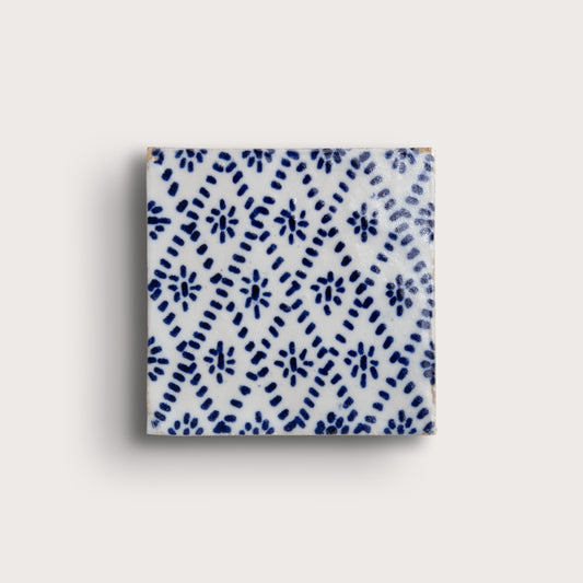 A square zellige tile with a white glaze and a blue hand-painted diamond pattern, shown on a light beige background.