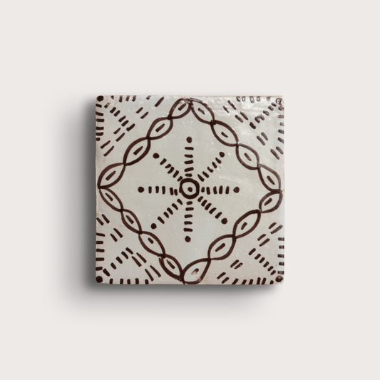 square zellige tile with a handpainted brown pattern shot on a light gray background.