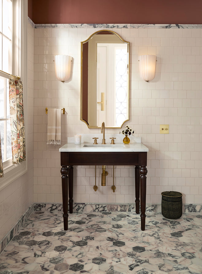 A vintage-style bathroom vanity with a dark wood pedestal sink, white marble countertop, gold faucet, and an arched gold-framed mirror. The room features white subway tiles, a dark reddish-brown upper wall, and a distinctive black, white, and gray marble-patterned floor.