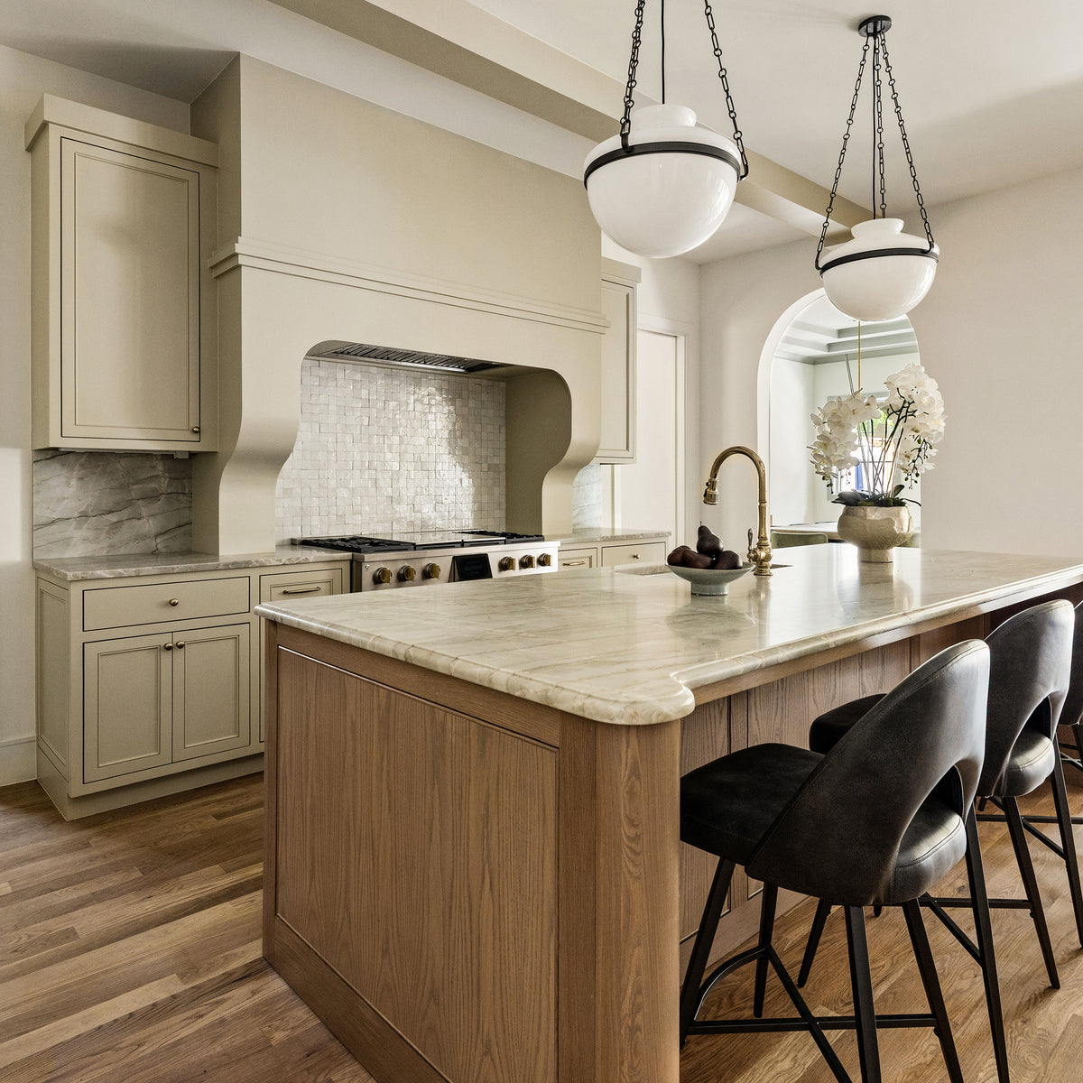 A modern kitchen island with a light wood base and a cream marble countertop, set in a spacious kitchen with beige cabinets, a tiled backsplash, and three black bar stools.