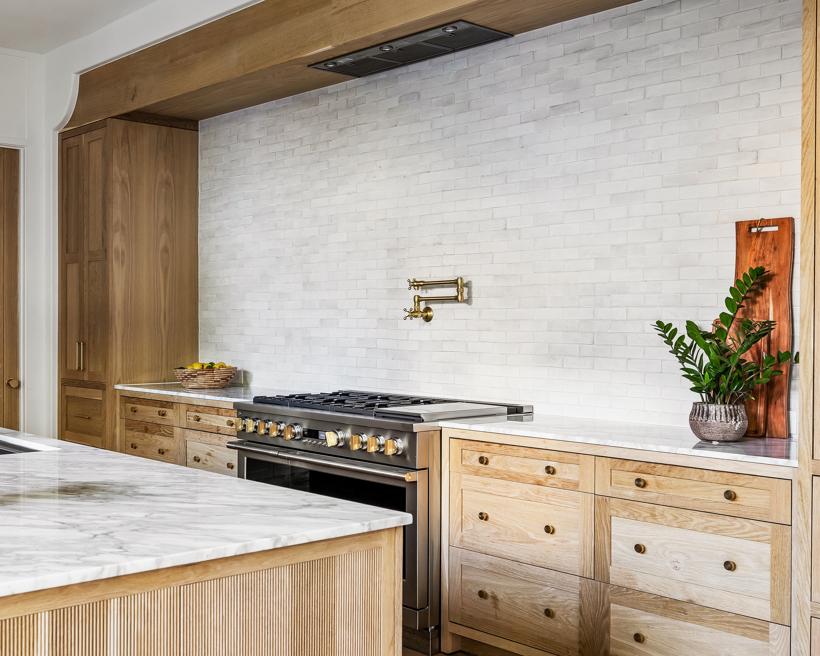 Modern kitchen with wooden cabinets, marble countertops, and white zellige tile as backsplash.