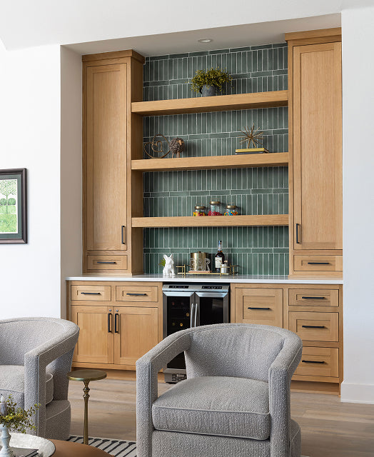 A built-in kitchen cabinet and shelving unit in a modern home interior, featuring light oak wood cabinetry, open shelves with decor, a green tile backsplash, a white countertop, and a built-in wine cooler. Two gray upholstered armchairs are partially visible in the foreground.