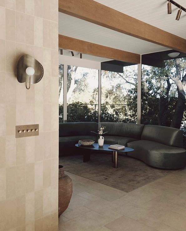 A modern curved sectional sofa in dark green leather sits in a sunlit contemporary home interior with large windows, wood beams, tiled walls, and a blue oval coffee table.