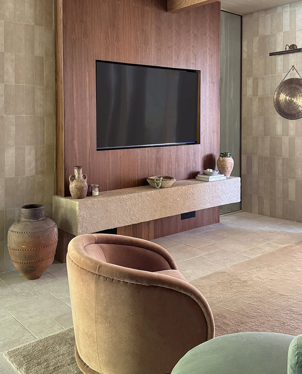 A modern living room interior featuring a large flat-screen TV mounted on a wood-paneled wall above a stone console shelf with decorative vases. A rounded blush-tan velvet armchair is prominent in the foreground, with a tiled wall and warm neutral decor throughout.