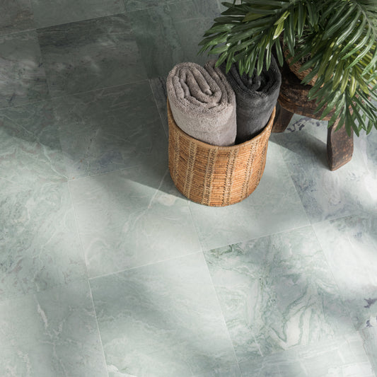 A styled interior scene showing a woven basket holding two rolled towels, one light gray and one dark charcoal, placed on a large pale green marble,floor tile next to a potted palm on a small wooden stool.