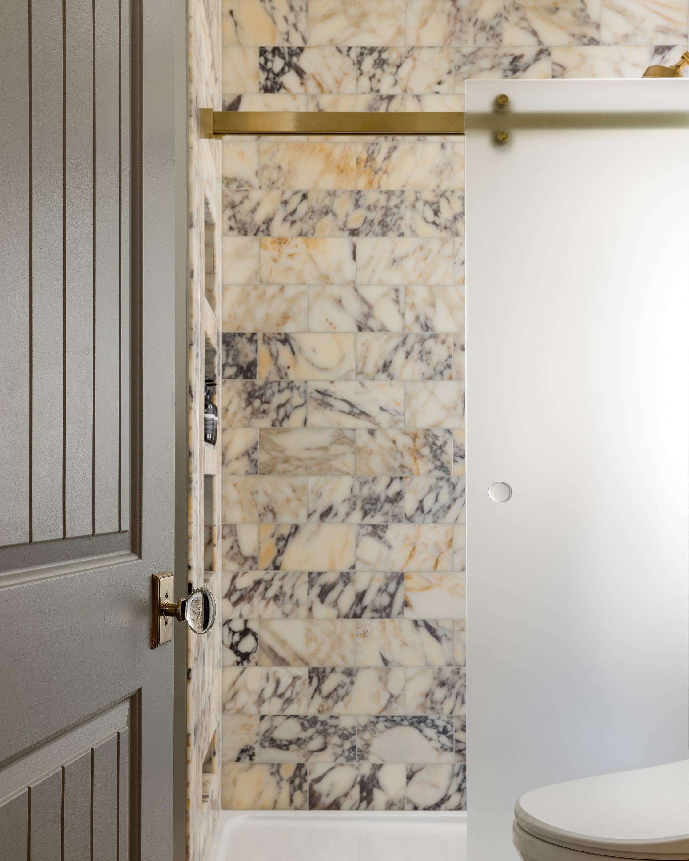 Marble-patterned shower wall with door and brass handle in a bathroom setting