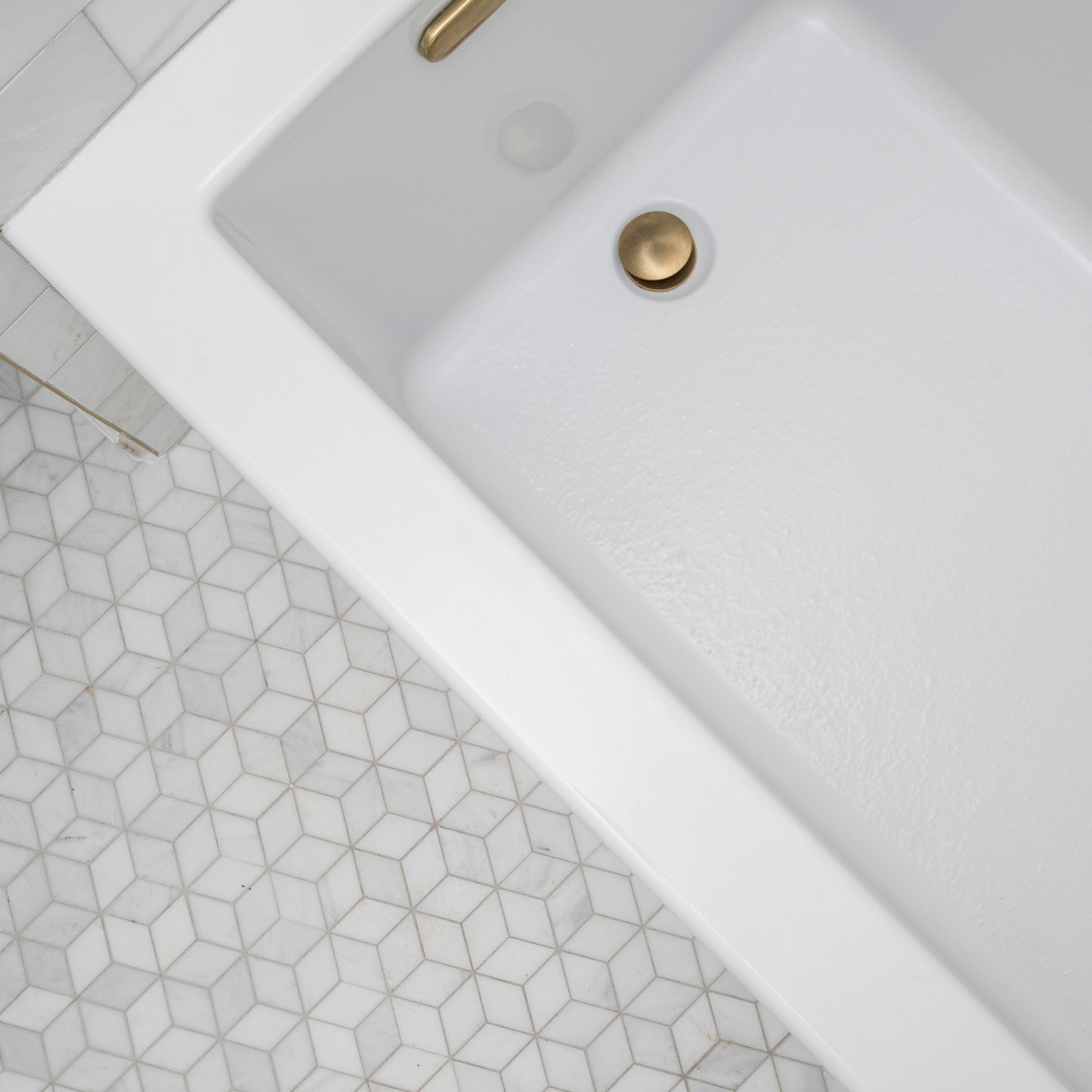 Close-up of a white kitchen sink with gold faucet and drain cover, geometric marble tile pattern on floor.