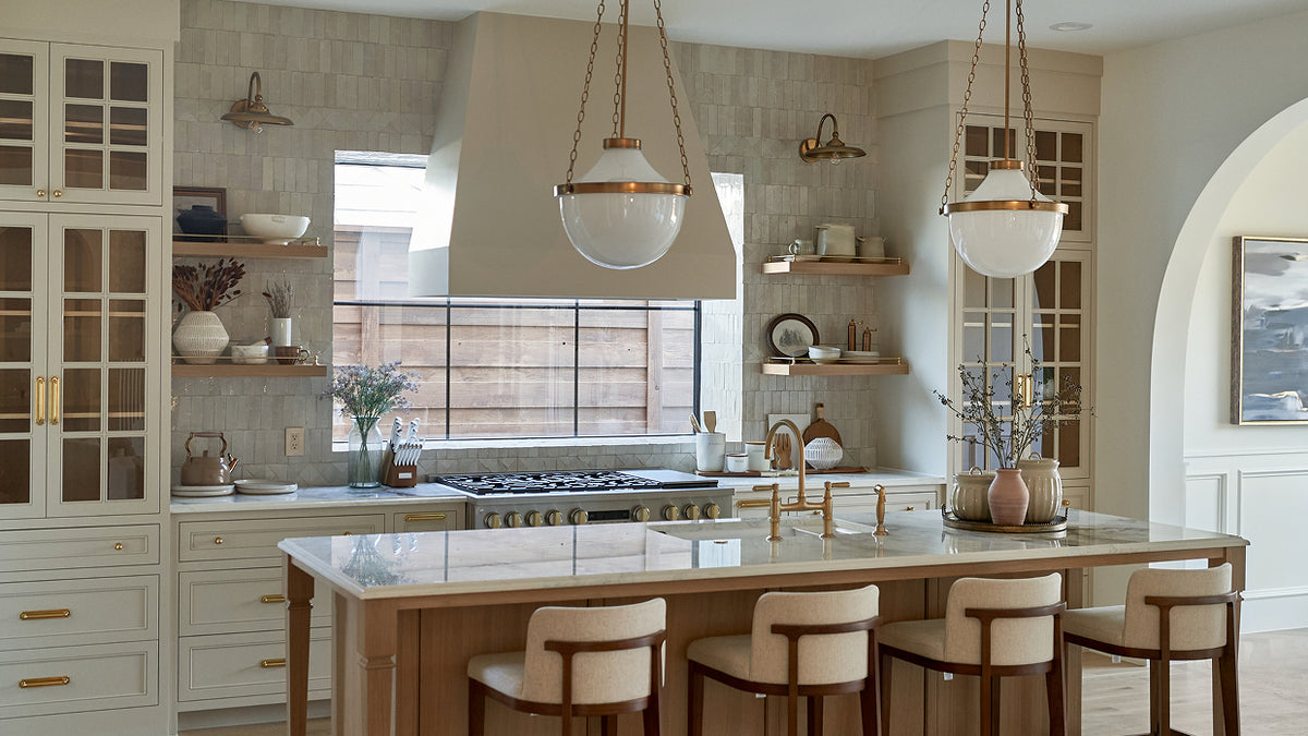 A bright, modern kitchen interior with cream cabinetry, a marble island, gold hardware, and two large pendant lights hanging above the counter.