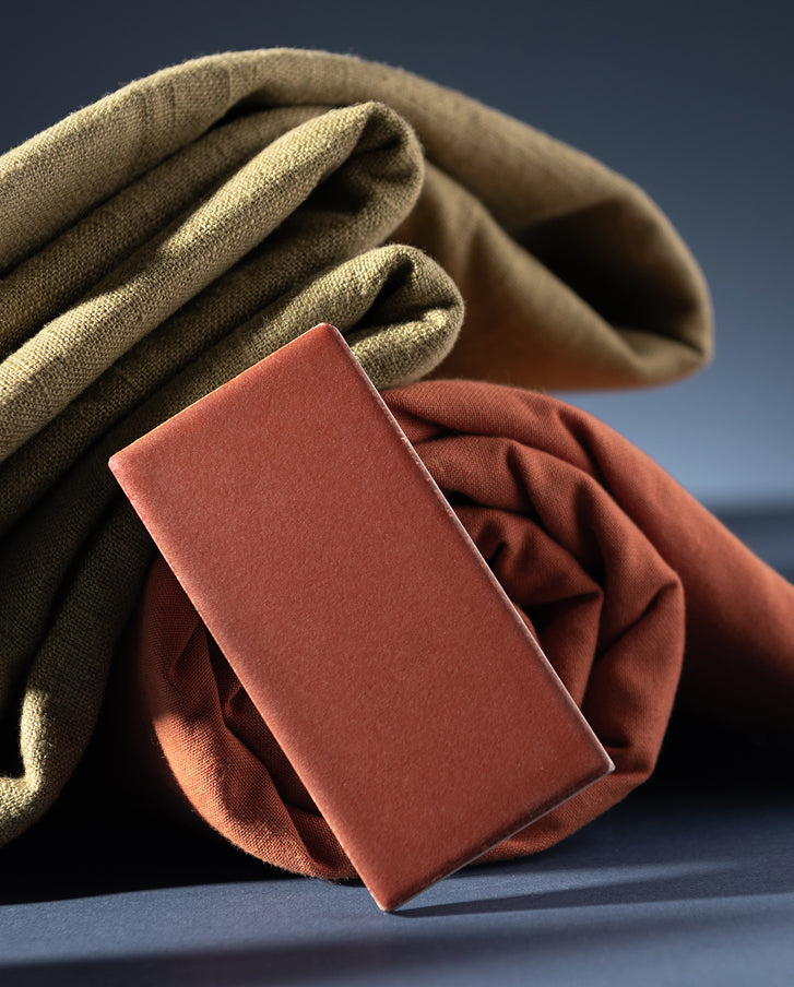Two folded fabric swatches, one in olive green and one in rust orange, arranged on a dark blue surface with a rectangular sample card showing the rust orange color in front.