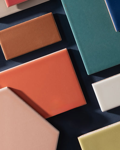 A styled flat-lay arrangement of various solid-colored rectangular tiles or swatches, including terracotta, coral, teal, white, pale pink, and pale yellow, set against a dark navy blue background.