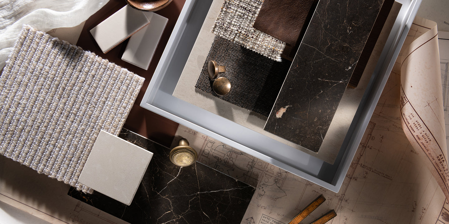 A styled flat-lay of home decor and material samples, featuring a textured woven runner, dark marble slabs, a metallic silver patterned runner, and a light blue tray on a beige map background.