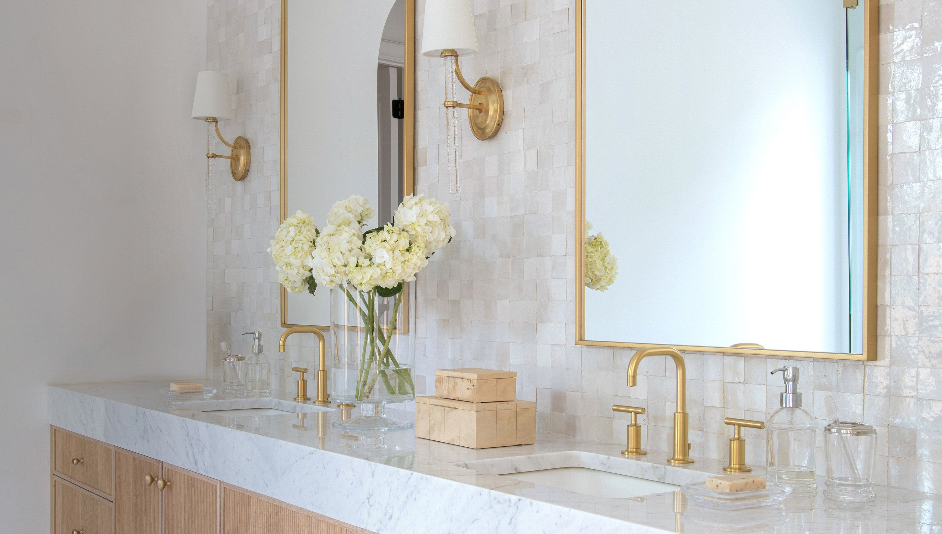 A luxury bathroom vanity featuring a white marble countertop, double sinks with polished gold faucets, a gold-framed mirror, and a vase of white flowers on a light wood cabinet.