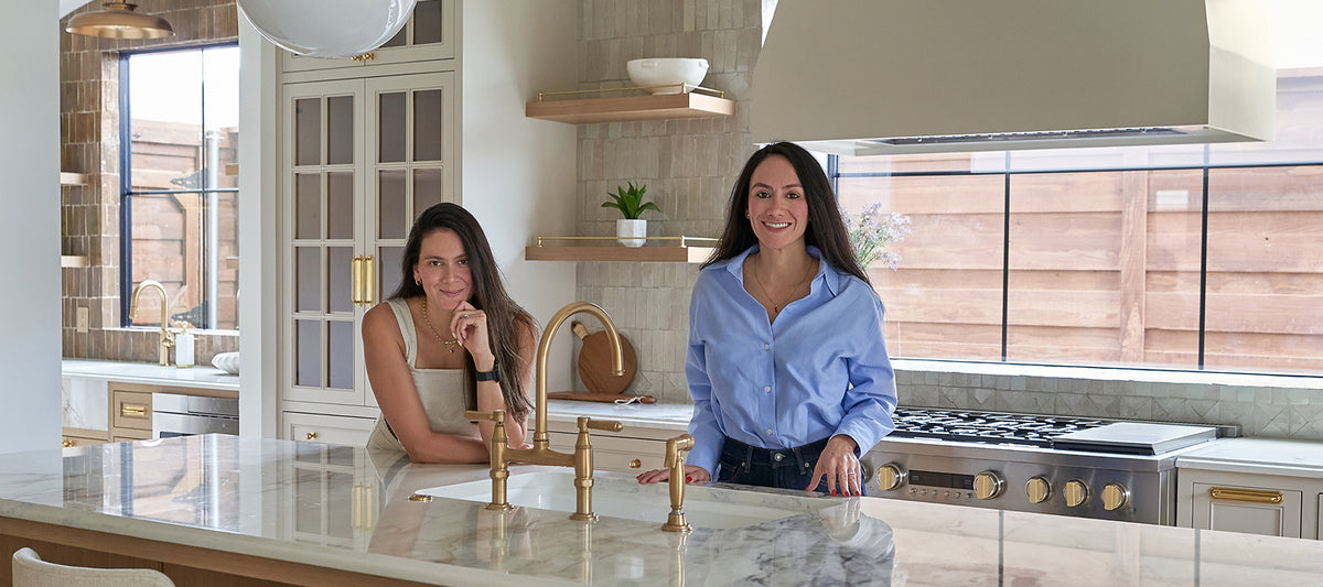Two women standing in a modern kitchen with white marble countertops, gold faucets, and light wood cabinetry.