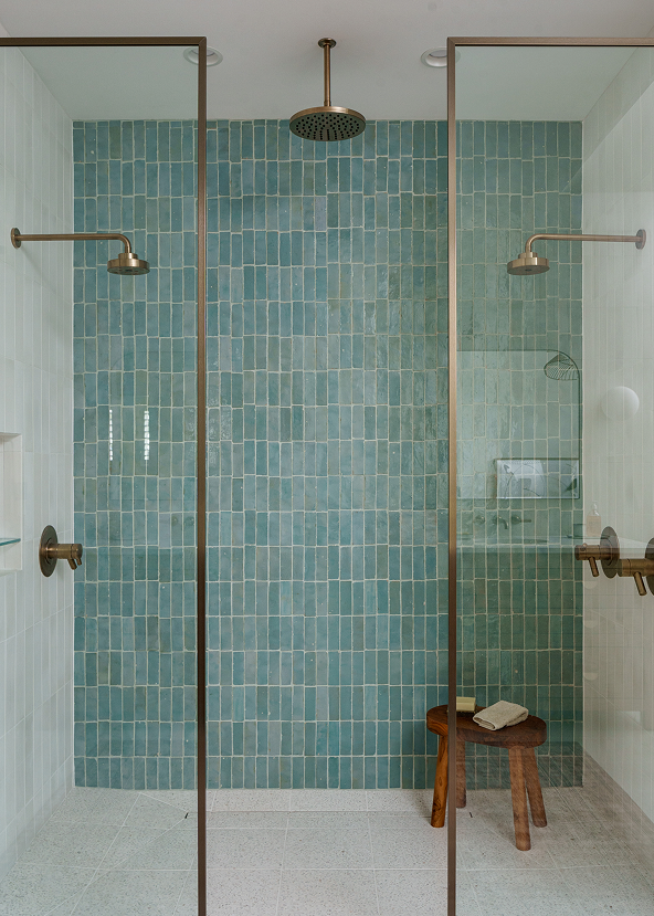 Modern bathroom with teal tiled zellige wall, glass shower enclosure, and wooden stool.