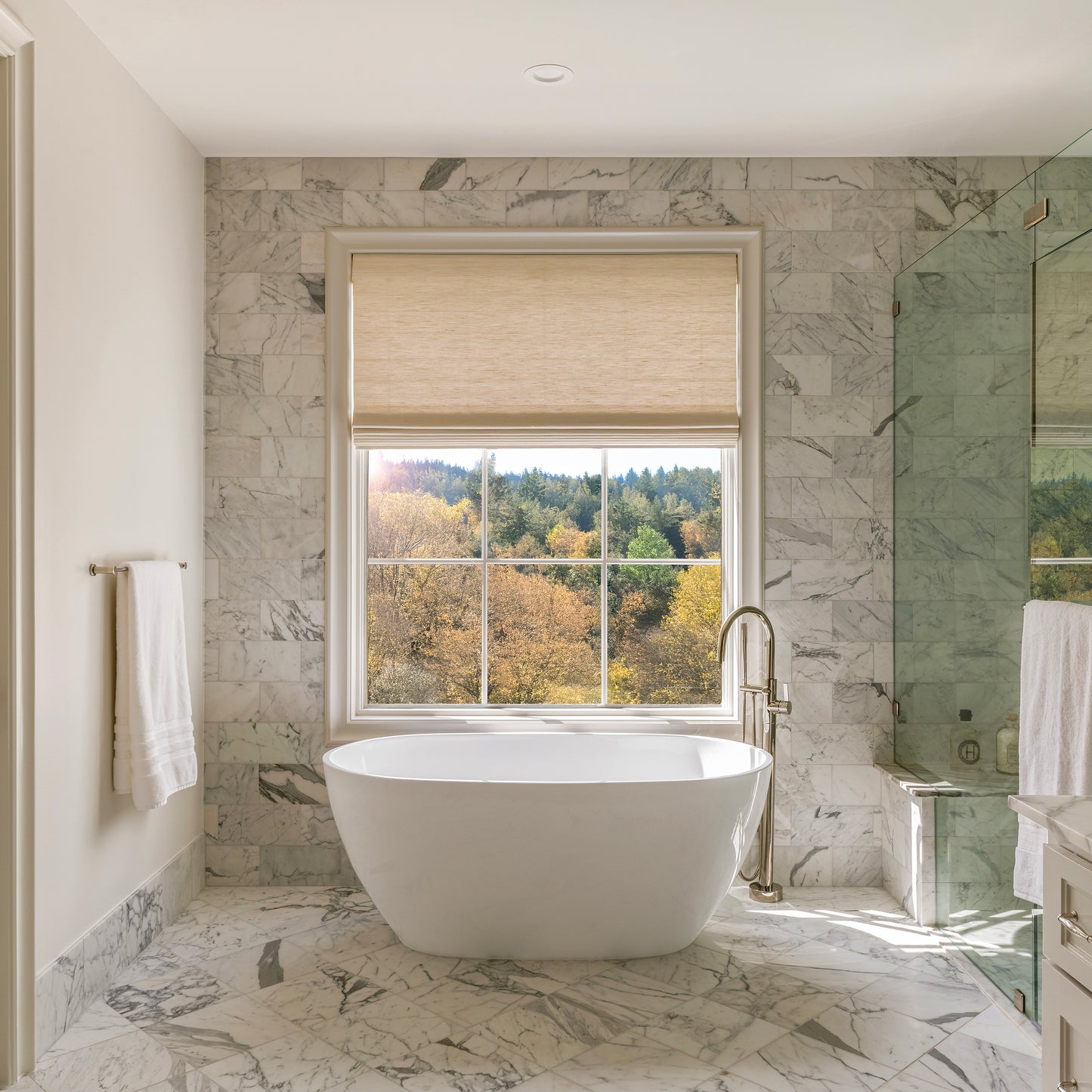 Modern bathroom with a freestanding bathtub, marble walls, and a window view of trees.
