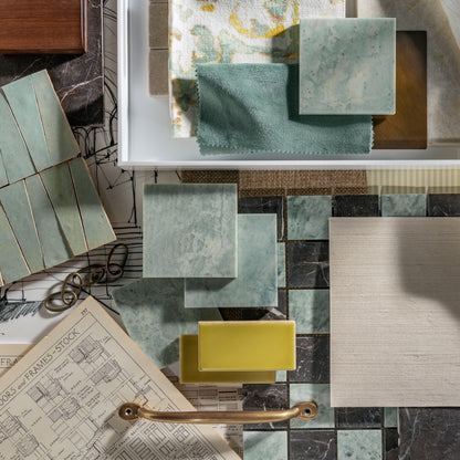 A styled flat-lay of home decor and marble and zellige materials, featuring square tiles and fabric and tile swatches in sage green, cream, and yellow, alongside a brass handle, architectural drawings, and wood textures.