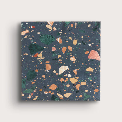 A square terrazzo-patterned tile or slab with a dark slate blue-gray base and scattered multicolored flecks in green, orange, peach, cream, and beige, shown against a light warm gray background.