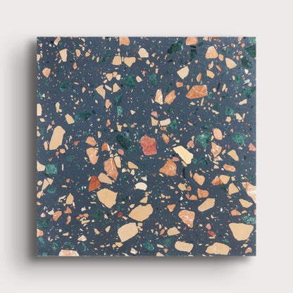 A square sample of terrazzo-style surface material with a dark blue-gray base and scattered multicolored chips in beige, orange, green, and white, shown against a light neutral background.