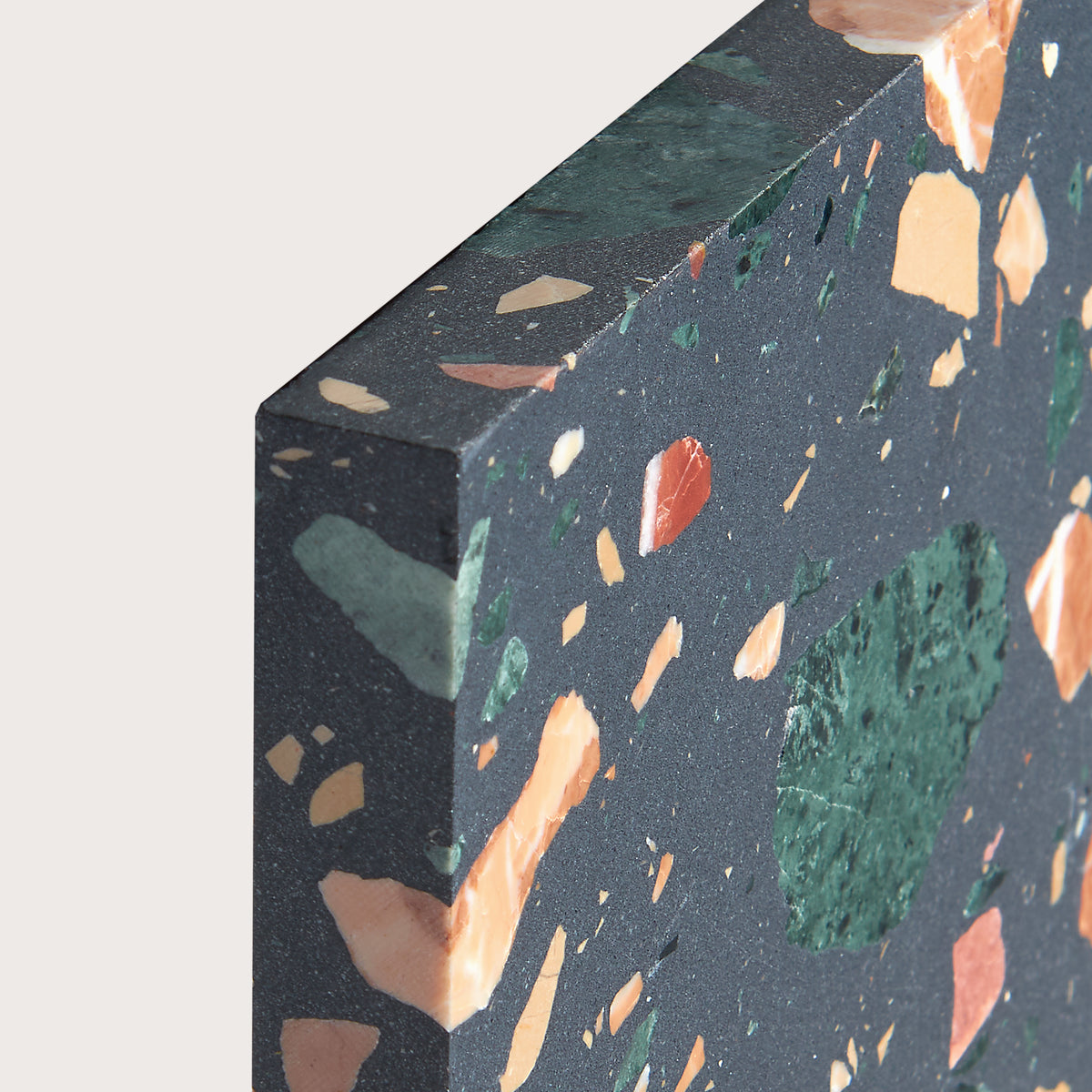 Close-up of a terrazzo slab showing a dark charcoal gray base with embedded orange, green, and peach flecks, photographed against a light neutral background.
