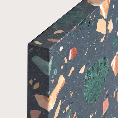 Close-up of a terrazzo slab showing a dark charcoal gray base with embedded orange, green, and peach flecks, photographed against a light neutral background.
