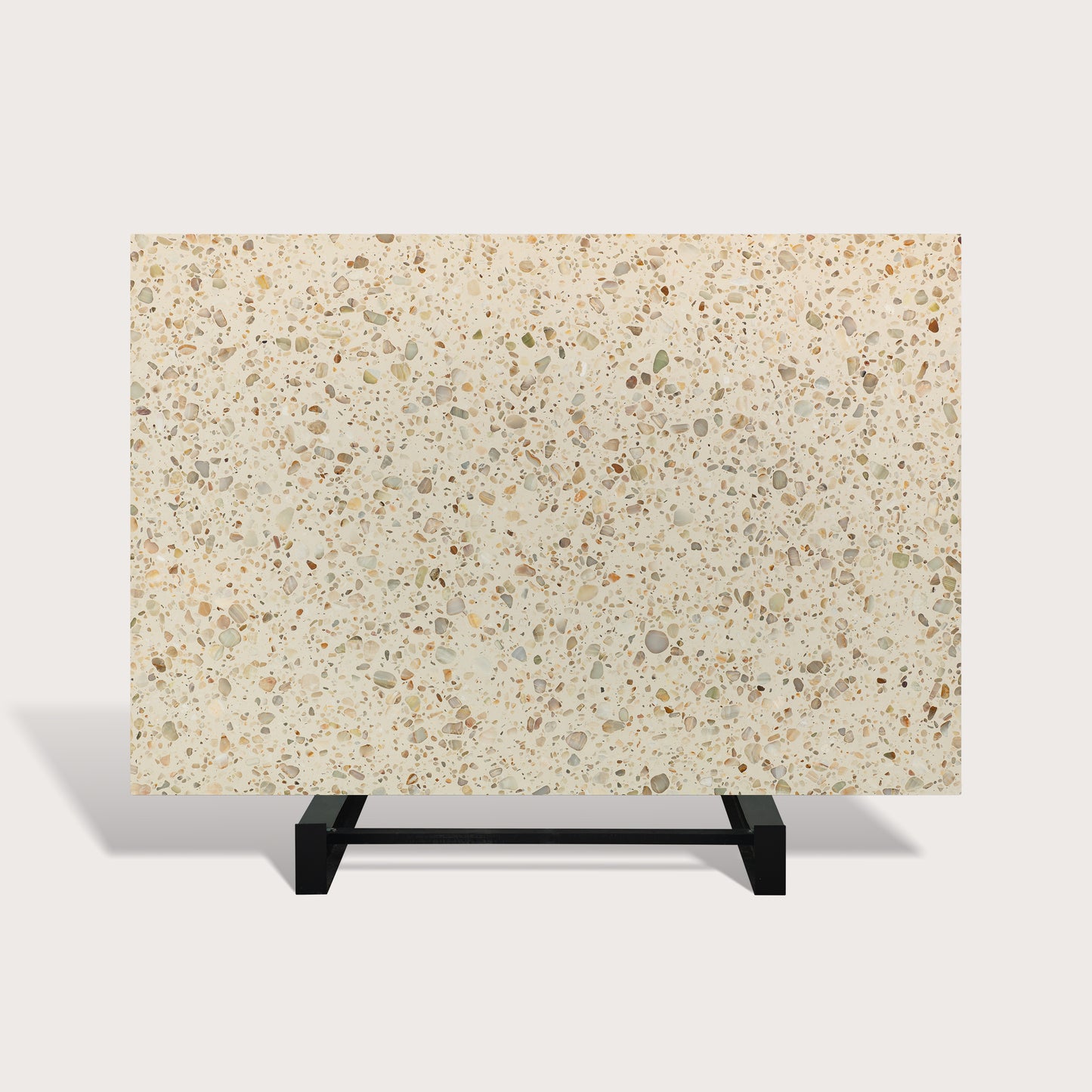 A rectangular terrazzo-style slab or tile sample with a warm cream background and scattered speckles in beige, gray, brown, and muted green, displayed on a black metal stand against a light gray studio background.