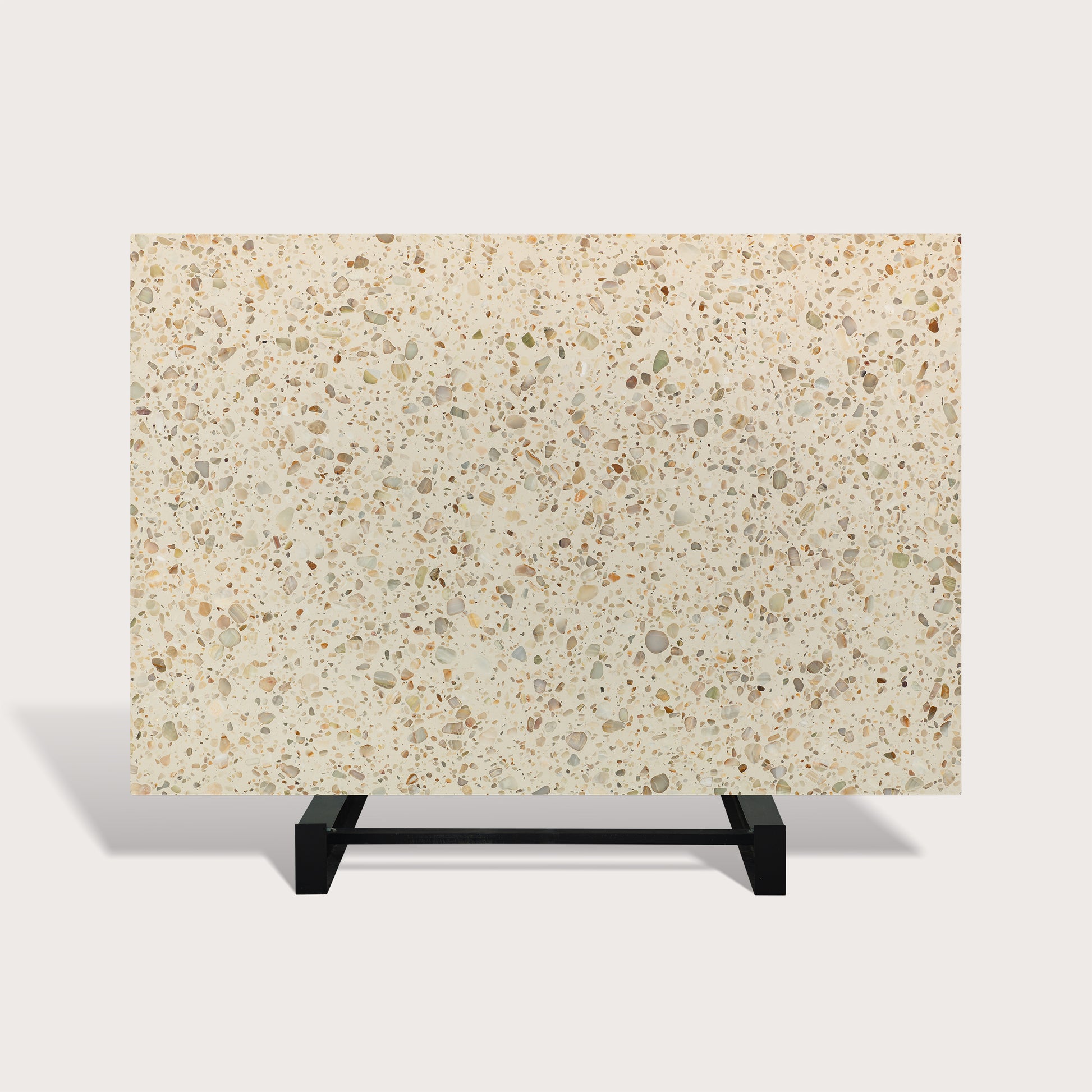 A rectangular terrazzo-style slab or tile sample with a warm cream background and scattered speckles in beige, gray, brown, and muted green, displayed on a black metal stand against a light gray studio background.