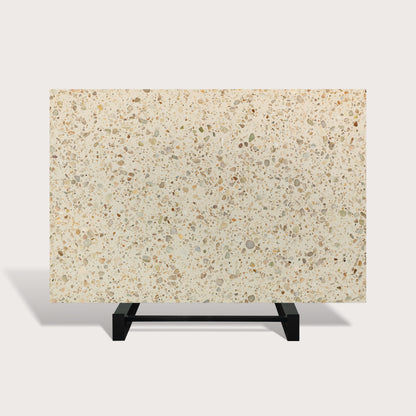 A rectangular terrazzo-style slab or tile sample with a warm cream background and scattered speckles in beige, gray, brown, and muted green, displayed on a black metal stand against a light gray studio background.