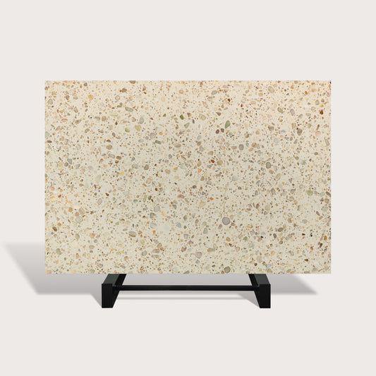 A rectangular terrazzo-style slab or tile sample with a warm cream background and scattered speckles in beige, gray, brown, and muted green, displayed on a black metal stand against a light gray studio background.