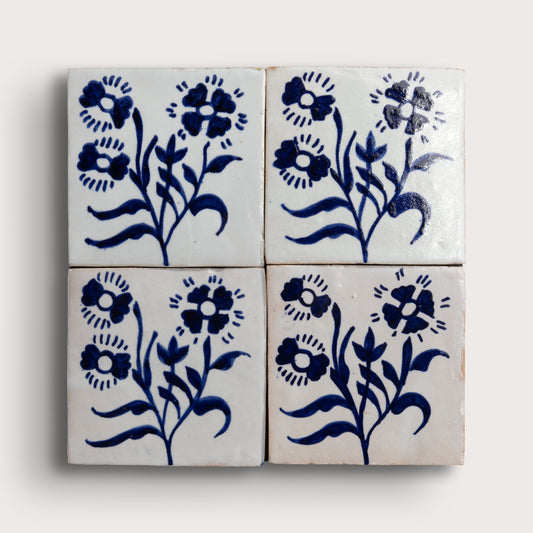Four square zellige tiles arranged in a 2x2 grid, featuring a hand-painted blue floral pattern on a glossy white glaze, set against a light beige background.