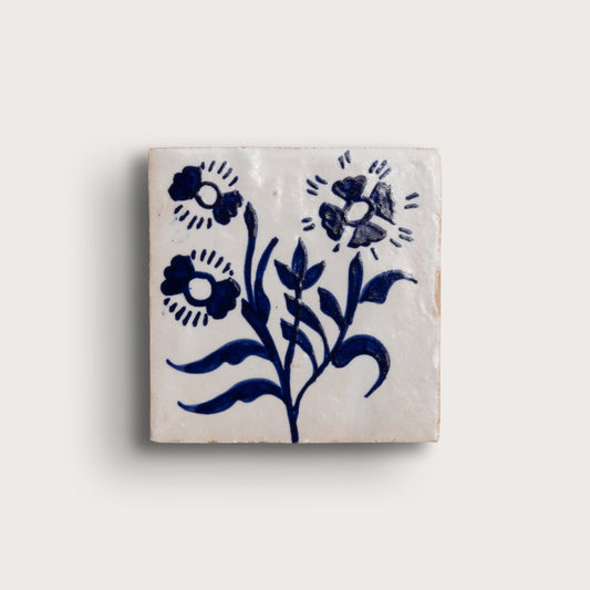A square zellige tile featuring a hand-painted blue floral design on a warm off-white background, shown against a solid light beige backdrop.