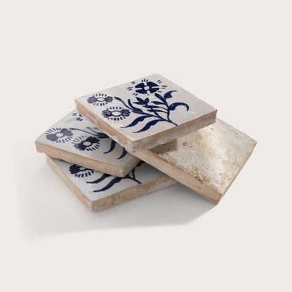Four square zellige tiles stacked in a pile on a light beige background. The tiles are off-white with a rustic, distressed finish and decorated with a dark blue floral pattern on the top tile.
