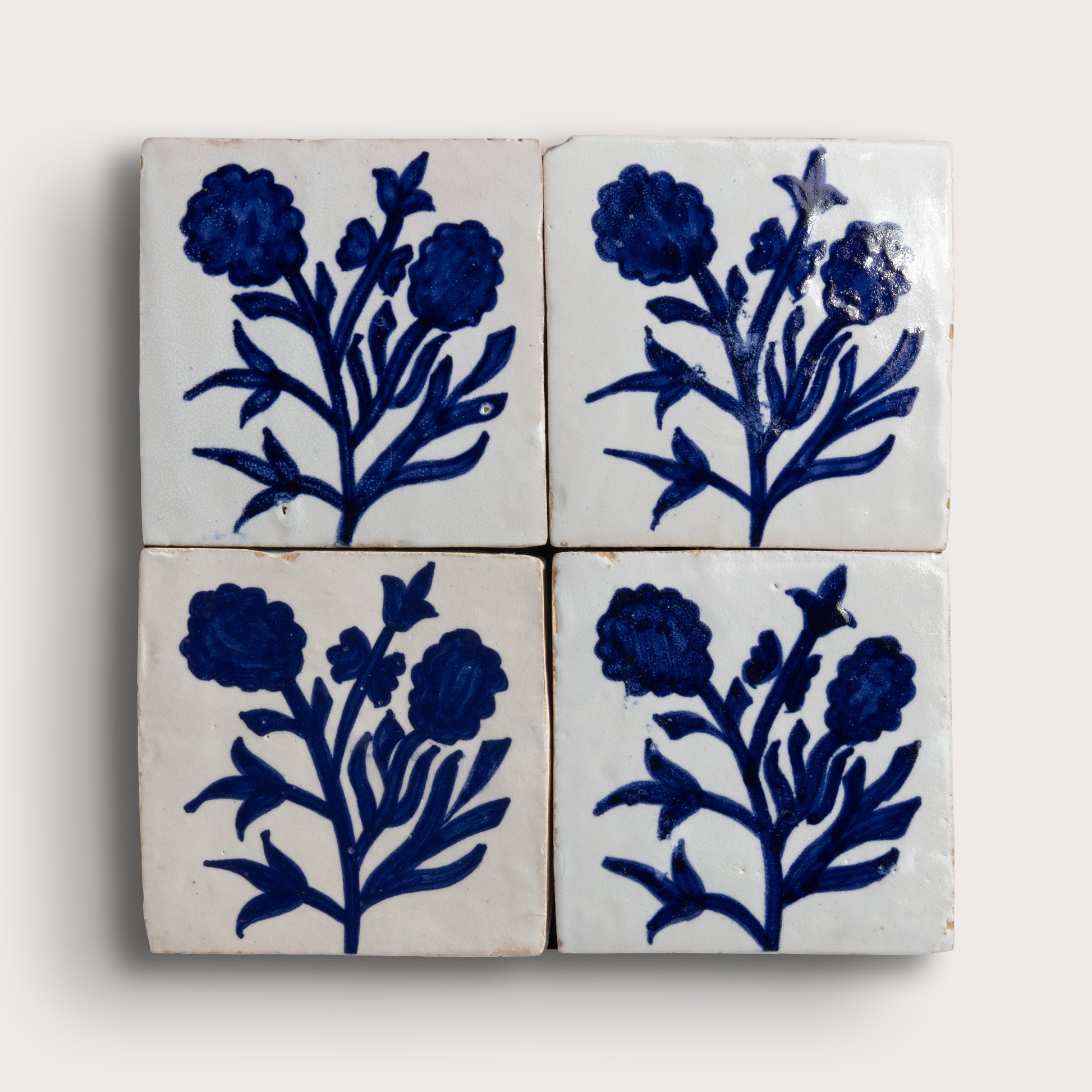 Four square zellige tiles arranged in a 2x2 grid, each decorated with a dark blue floral motif on an off-white background, photographed against a light neutral backdrop.