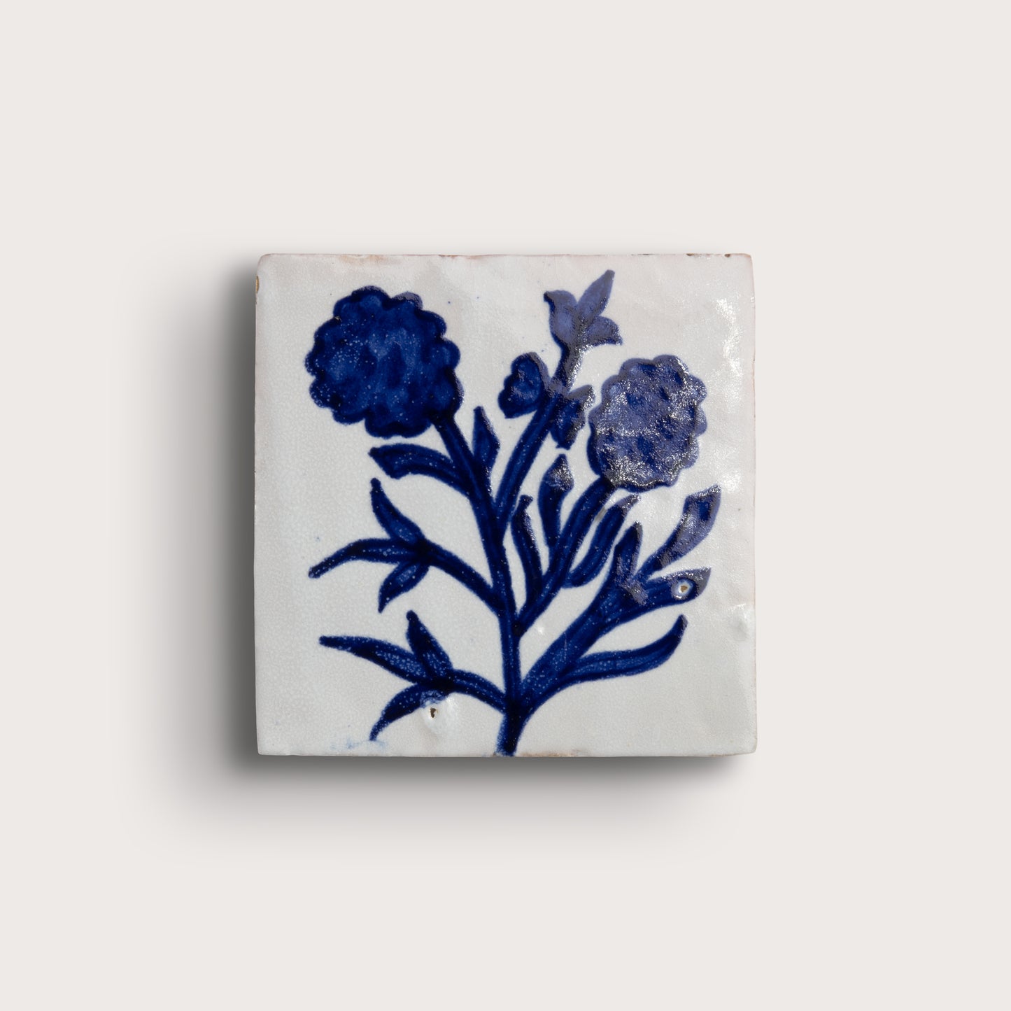 A square white zellige tile featuring a hand-painted blue floral design, displayed against a solid off-white background.