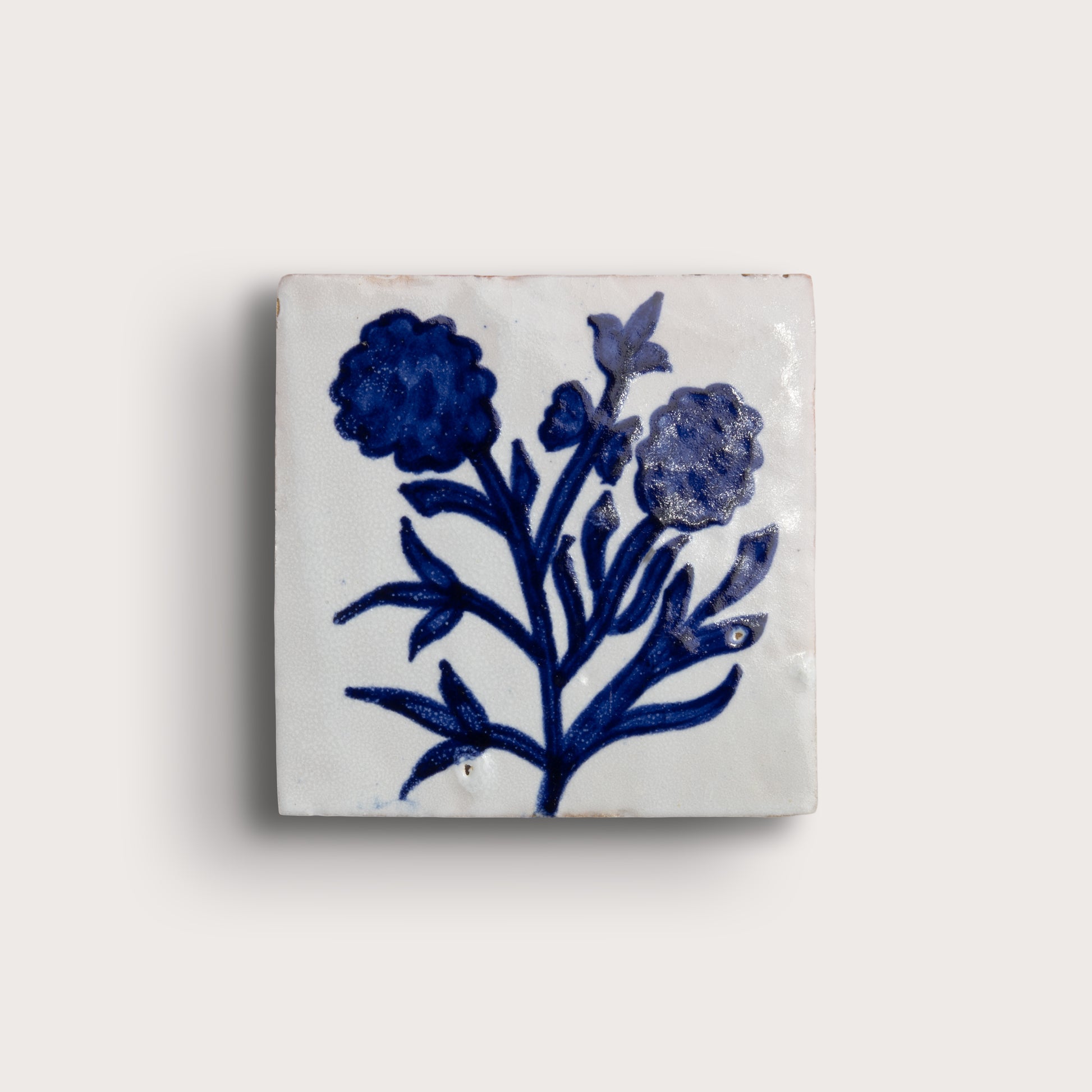 A square white zellige tile featuring a hand-painted blue floral design, displayed against a solid off-white background.