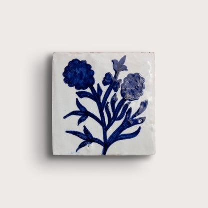 A square white zellige tile featuring a hand-painted blue floral design, displayed against a solid off-white background.
