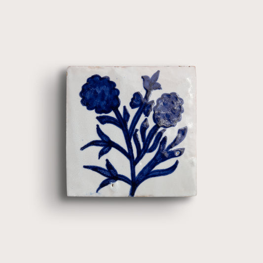 A square white zellige tile featuring a hand-painted blue floral design, displayed against a solid off-white background.