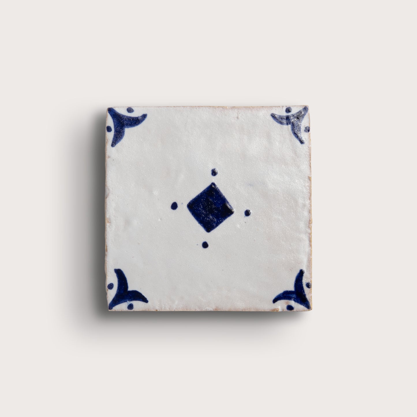 A square zellige tile with a white glaze and dark blue hand-painted motifs, featuring a central diamond and corner flourishes, shown on a light beige background.