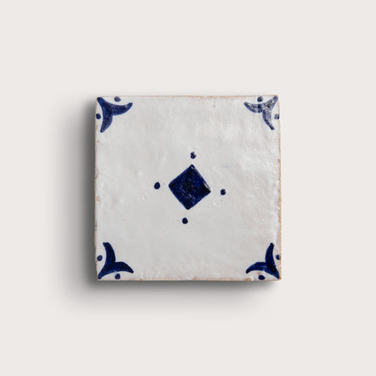 A square zellige tile with a white glaze and dark blue hand-painted motifs, featuring a central diamond and corner flourishes, shown on a light beige background.