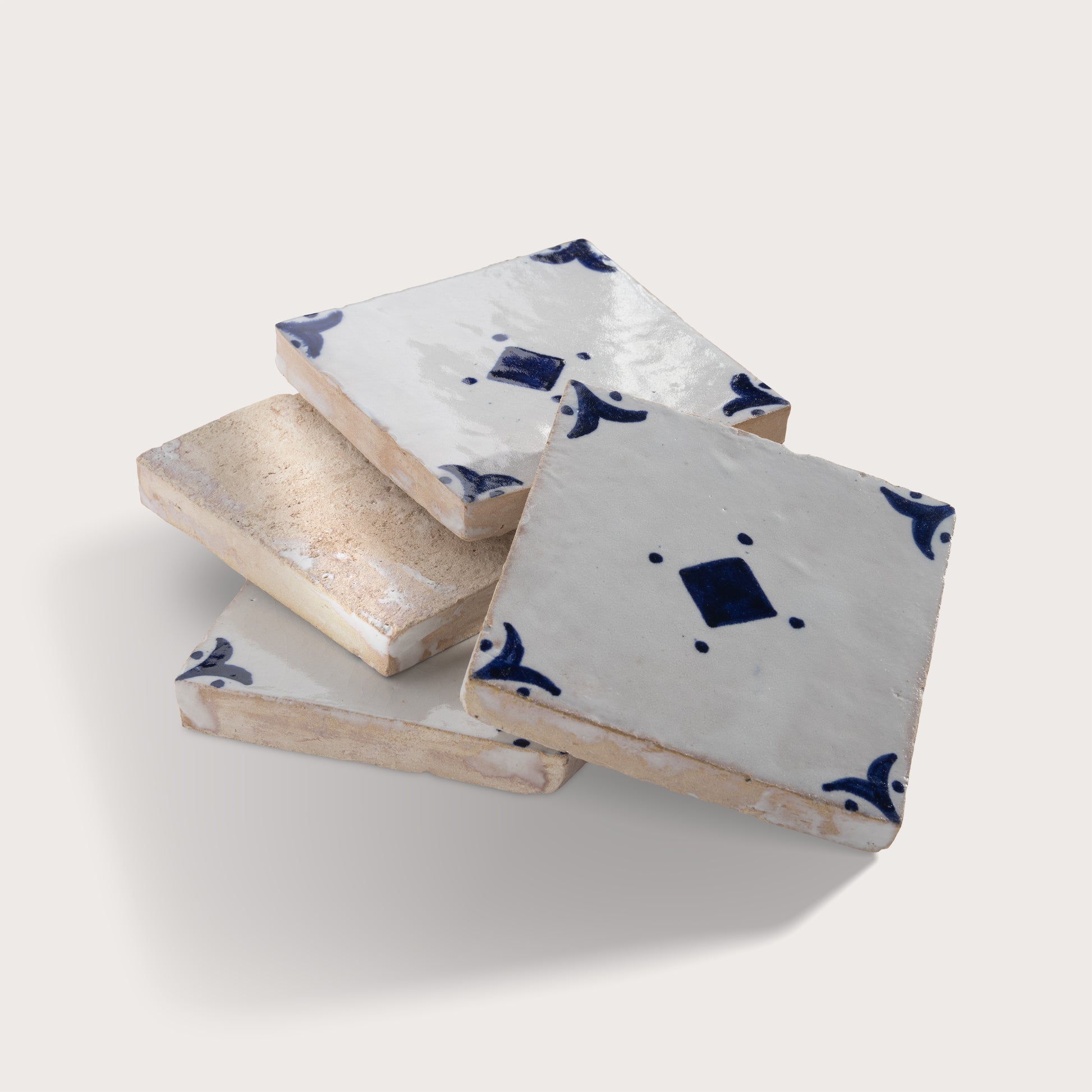 Four square zellige tiles stacked and overlapping on a light beige background. Three tiles are glazed in white with dark blue hand-painted traditional patterns, while one tile has a rough, unglazed beige surface.