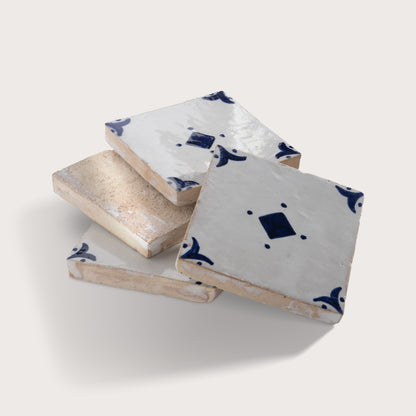 Four square zellige tiles stacked and overlapping on a light beige background. Three tiles are glazed in white with dark blue hand-painted traditional patterns, while one tile has a rough, unglazed beige surface.