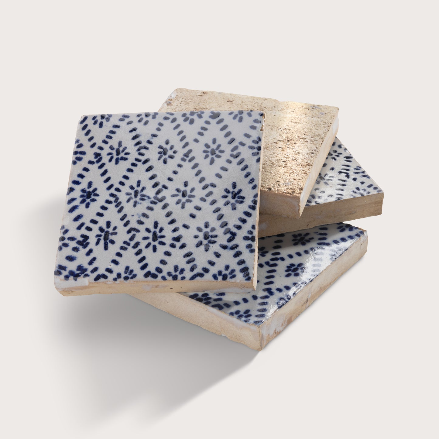 Three square zellige tiles stacked in a fan arrangement on a light beige background. The top tile is white with a dark blue hand-painted floral and dotted pattern, while the bottom two tiles show a textured, unglazed beige clay side.