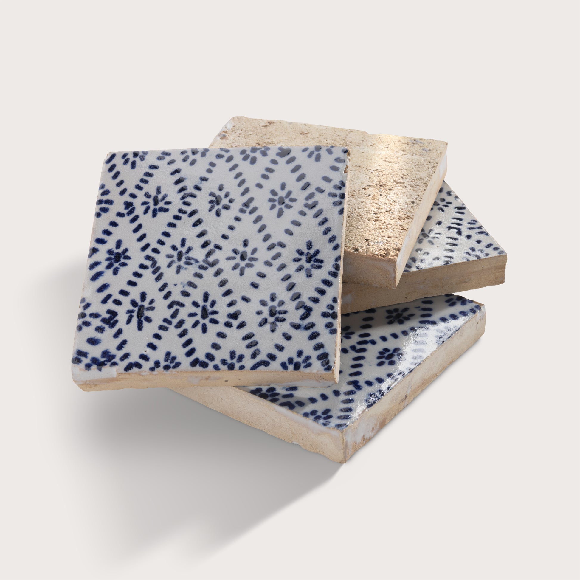 Three square zellige tiles stacked in a fan arrangement on a light beige background. The top tile is white with a dark blue hand-painted floral and dotted pattern, while the bottom two tiles show a textured, unglazed beige clay side.