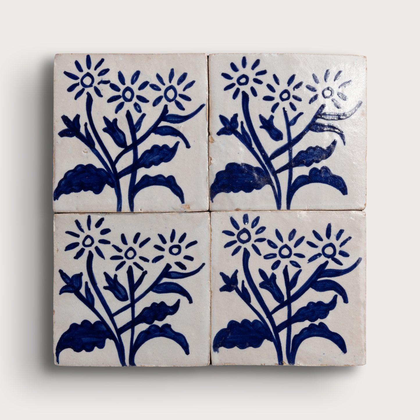 Four square zellige tiles arranged in a 2x2 grid, each decorated with a hand-painted blue floral motif on an off-white background, set against a light neutral backdrop.