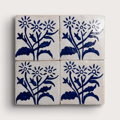 Four square zellige tiles arranged in a 2x2 grid, each decorated with a hand-painted blue floral motif on an off-white background, set against a light neutral backdrop.