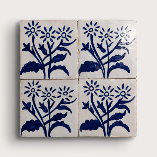 Four square zellige tiles arranged in a 2x2 grid, each decorated with a hand-painted blue floral motif on an off-white background, set against a light neutral backdrop.