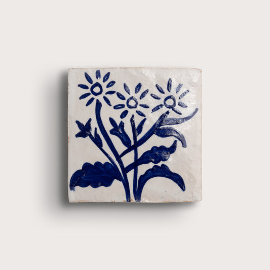 A square zellige tile featuring a hand-painted blue floral design on a warm off-white background, shown against a solid light beige backdrop.