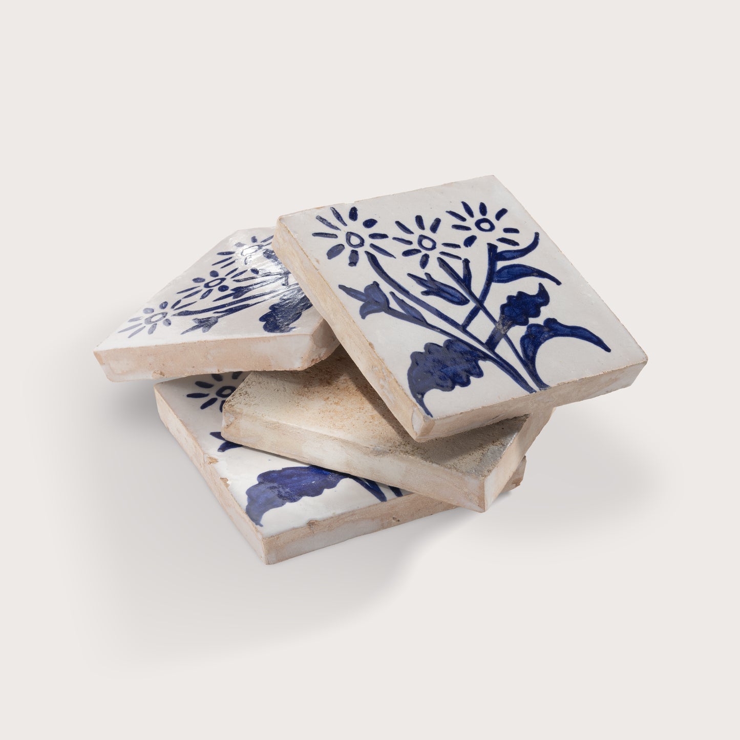 Four square zellige tiles stacked in a pile, featuring a blue floral and botanical pattern on a warm off-white background, set against a solid light beige backdrop.