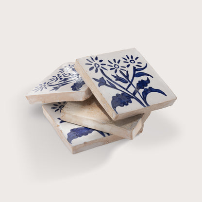 Four square zellige tiles stacked in a pile, featuring a blue floral and botanical pattern on a warm off-white background, set against a solid light beige backdrop.