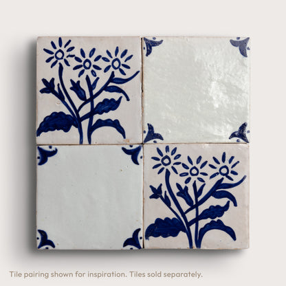 Four square zellige tiles arranged in a 2x2 grid, featuring a classic blue and white floral pattern on two tiles and solid white glaze on the other two, set against a light beige background.